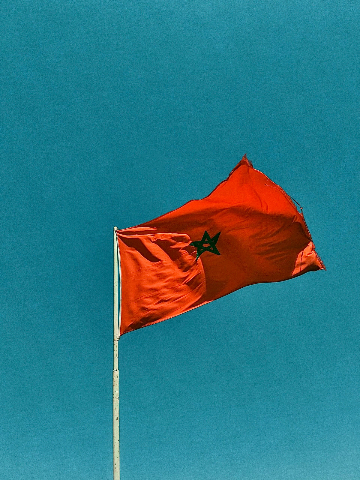 Drapeau rouge du Maroc avec une étoile verte au centre, flottant au sommet d’un mât sur un ciel bleu.