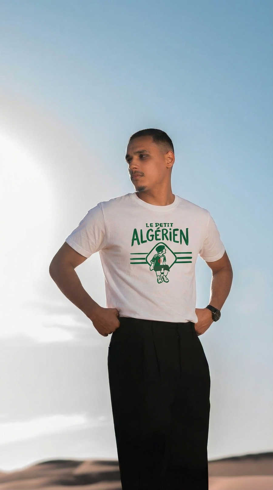 Vue de face du t-shirt blanc Le Petit Algérien, porté avec un pantalon noir devant un décor de dunes.