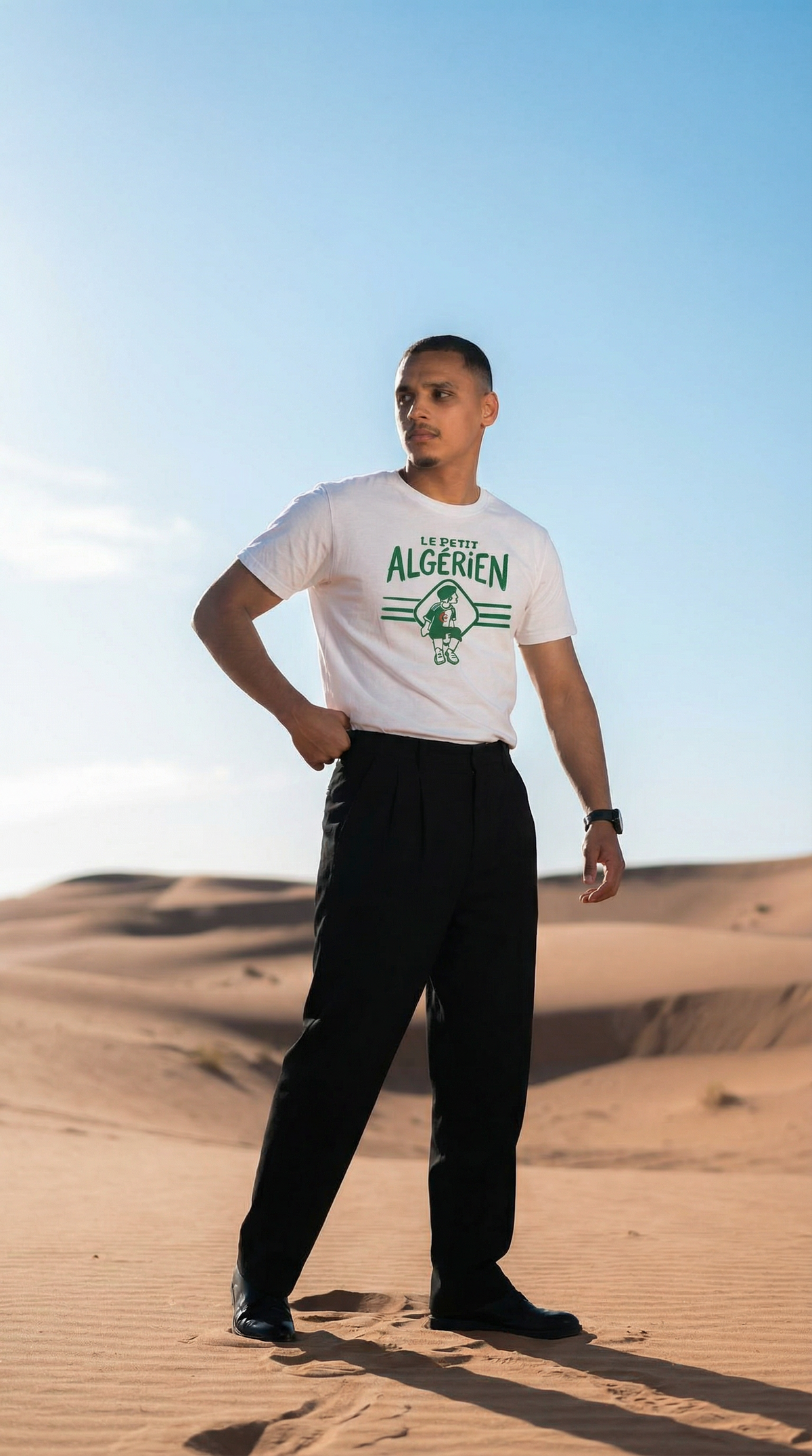 T-shirt Le Petit Algérien porté en pied dans les dunes, avec un pantalon noir et des chaussures sombres.